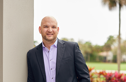 Man in a dark suit with a lavender shirt leaning against a beige column outdoors, with greenery in the background.