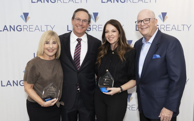 Four smiling adults posing at an awards event, two women holding glass trophies in front of a Lang Realty backdrop.